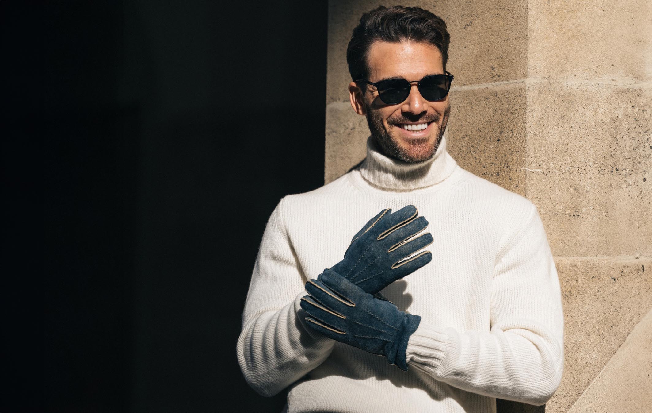 Mann mit weissem Rollkragenpullover und handgefertigten blauen Lederhandschuhen von Thomas Riemer Handschuhmanufaktur in Wien, lächelnd vor einer Steinwand.