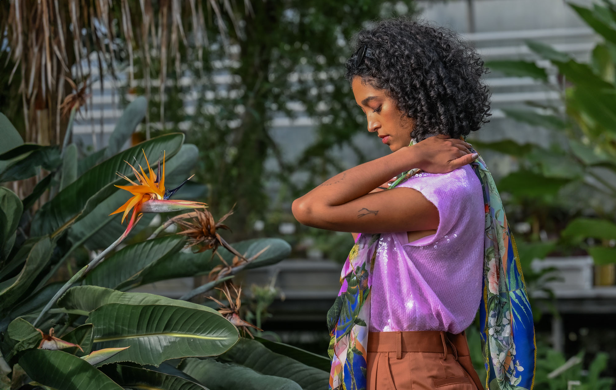Eine Frau mit lockigem Haar trägt ein fliederfarbenes Paillettentop und einen bunten Schal mit Naturmotiven, während sie sich sanft an der Schulter berührt. Im Vordergrund blüht eine auffällige Paradiesvogelblume.