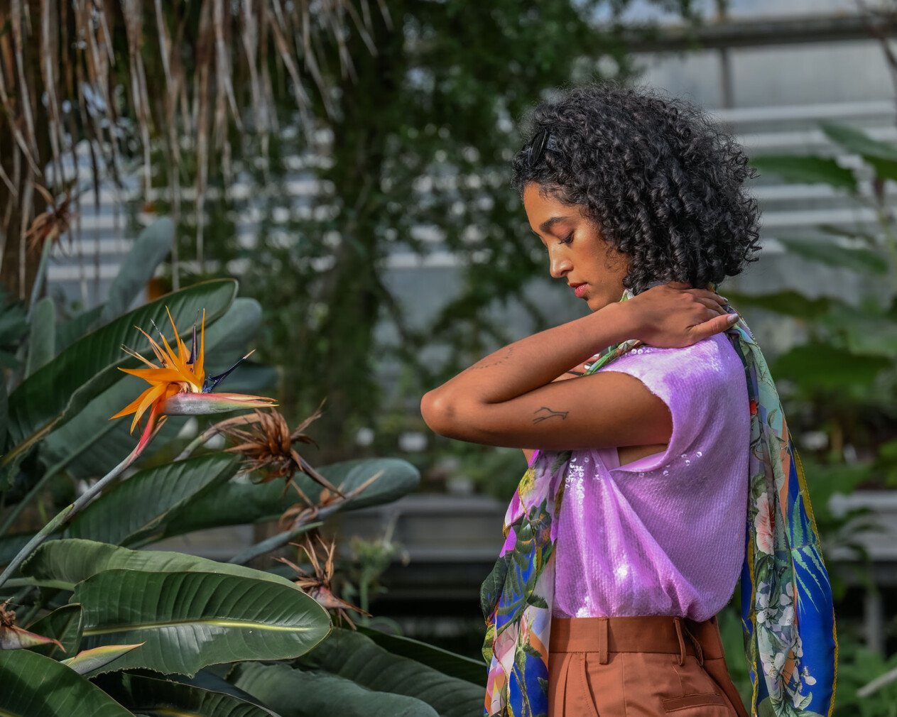 Eine Frau mit lockigem Haar trägt ein fliederfarbenes Paillettentop und einen bunten Schal mit Naturmotiven, während sie sich sanft an der Schulter berührt. Im Vordergrund blüht eine auffällige Paradiesvogelblume.