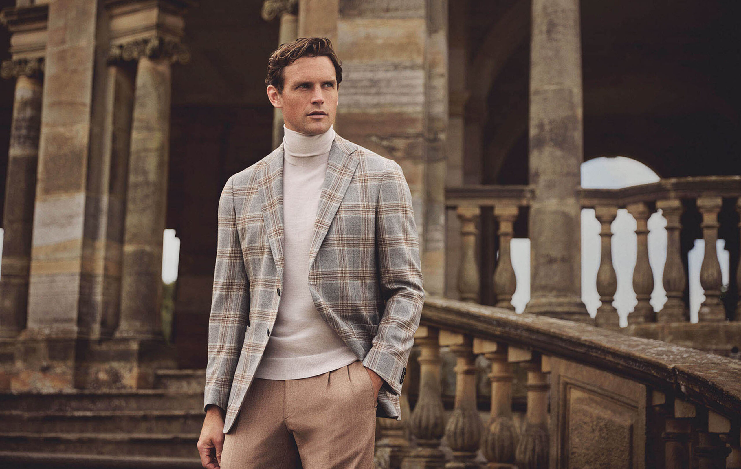 scabal-herrenbekleidung-herbst-2024 Elegantes Herbstoutfit von Scabal: Ein Mann trägt ein kariertes Sakko in warmen Erdtönen mit Rollkragenpullover und Stoffhose vor historischer Kulisse.