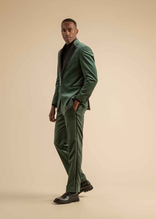 Dreiteiliger Herrenanzug aus grünem Samt mit schwarzem Revers, kombiniert mit schwarzem Rollkragenpullover, getragen von einem Model vor beigem Hintergrund.