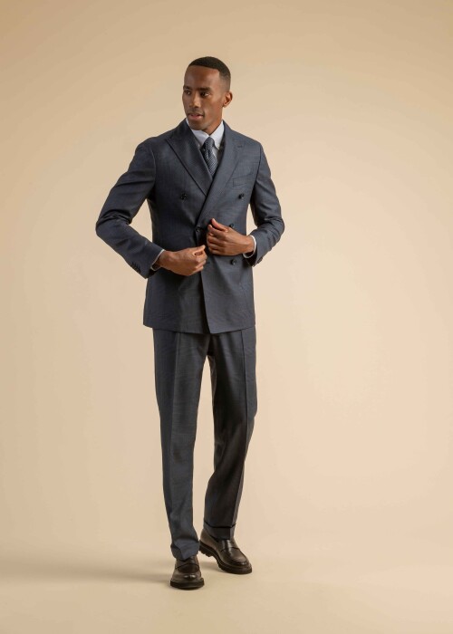 Herrenanzug in dunkelblauem Stoff mit feinem Karomuster und zweireihigem Sakko, kombiniert mit Hemd und Krawatte, getragen von einem Model vor beigem Hintergrund.