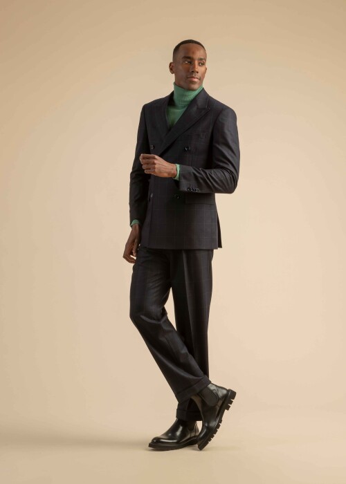 Herrenanzug in dunkelblauem Karomuster mit zweireihigem Sakko, kombiniert mit grünem Rollkragenpullover und schwarzen Chelsea Boots, getragen von einem Model vor beigem Hintergrund.