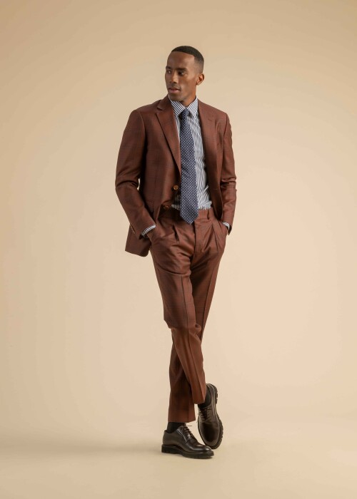Herrenanzug in rostbraunem Karomuster, kombiniert mit gestreiftem Hemd und dunkelblauer Krawatte mit Punkten, getragen von einem Model vor beigem Hintergrund.