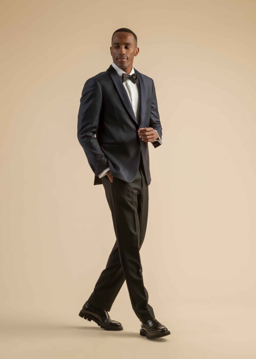 Dunkelblauer Smoking mit schwarzem Revers aus der Scabal Herbstkollektion Abendanzug mit dunkelblauem Jackett, schwarzem Revers, weissem Hemd und schwarzer Fliege, kombiniert mit schwarzer Hose und schwarzen Lackschuhen, getragen von einem Model vor beigem Hintergrund.