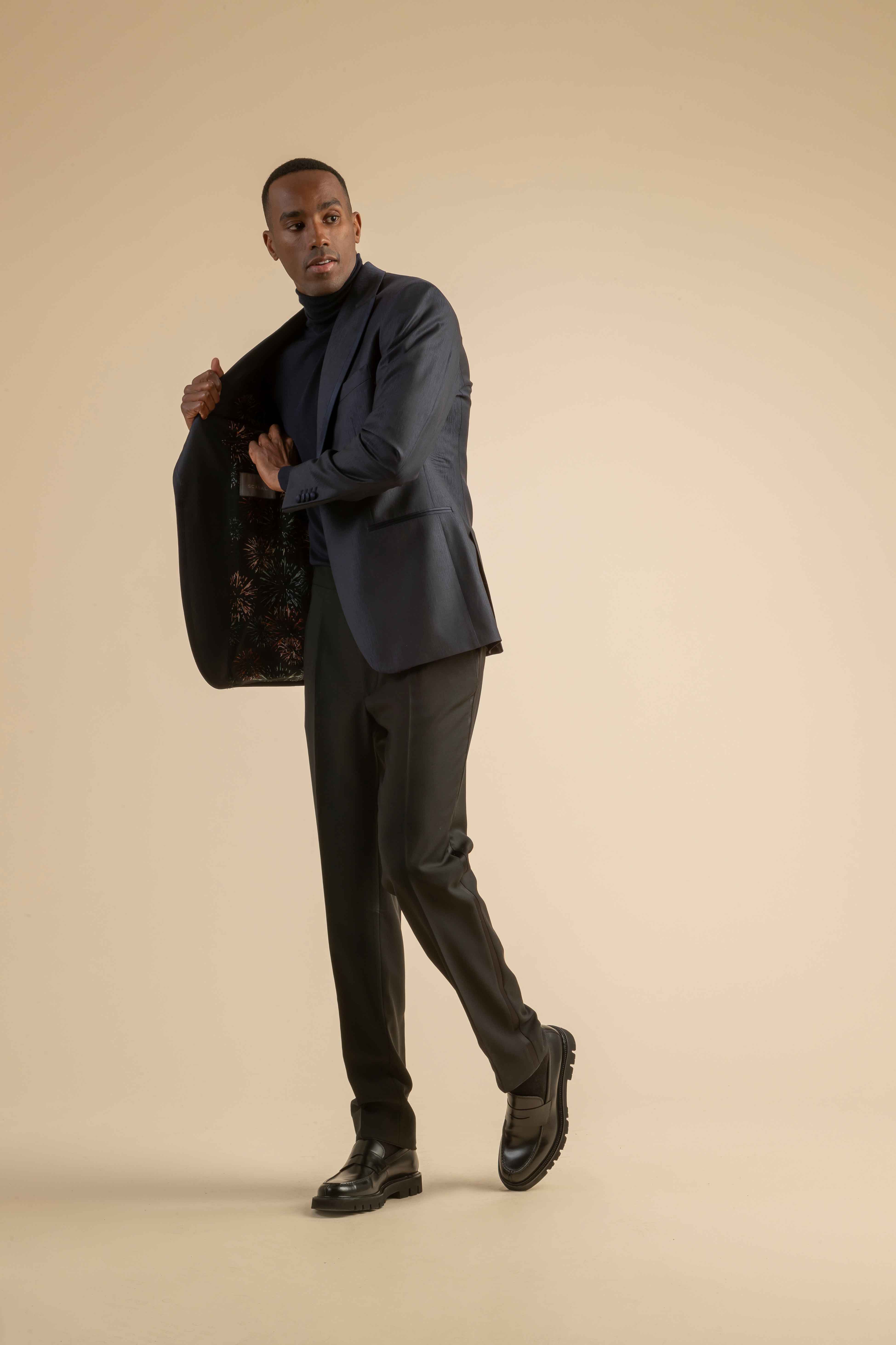 Dunkelblauer Anzug mit gemustertem Innenfutter aus der Scabal Herbstkollektion Dunkelblauer Herrenanzug mit schwarzem Rollkragenpullover, geöffnetem Jackett mit gemustertem Innenfutter, getragen von einem Model vor beigem Hintergrund.