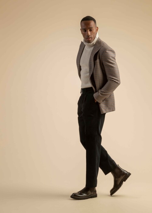 Kariertes Sakko mit Rollkragenpullover aus der Scabal Herbstkollektion Herrenoutfit mit braun-blau kariertem Sakko, grob gestricktem weissem Rollkragenpullover und dunkler Stoffhose, getragen von einem Model vor beigem Hintergrund.