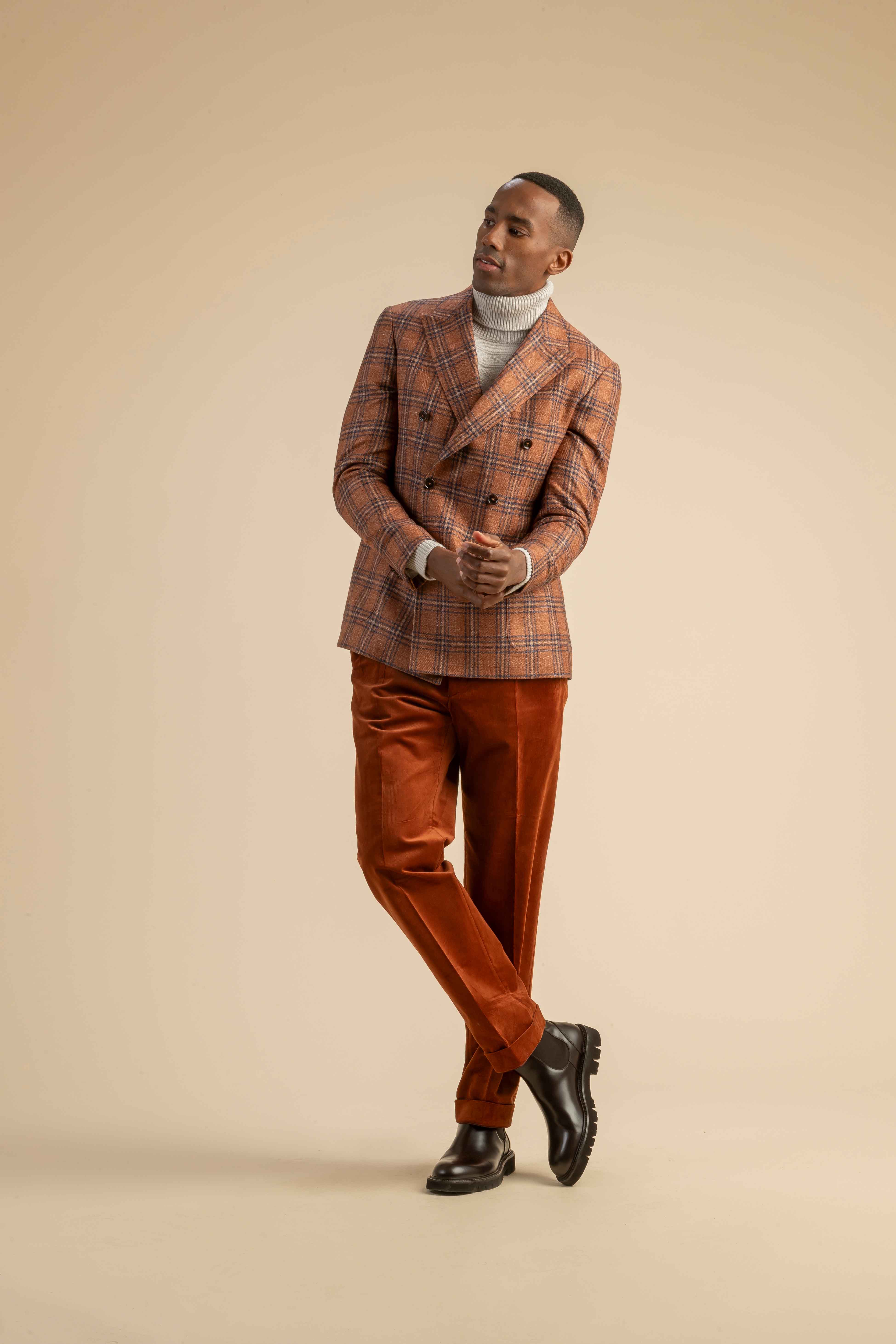 Kariertes Sakko mit Cordhose aus der Scabal Herbstkollektion Herrenoutfit mit orange-braun kariertem Doppelreiher-Sakko, beigefarbenem Rollkragenpullover und rostroter Cordhose, getragen von einem Model vor beigem Hintergrund.