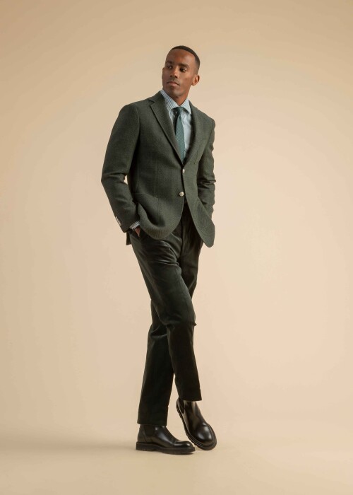 Grünes Sakko mit Hemd und Cordhose aus der Scabal Herbstkollektion Herrenoutfit mit grünem Wollsakko, grün gestreiftem Hemd, dunkler Krawatte und dunkelgrüner Cordhose, getragen von einem Model vor beigem Hintergrund.