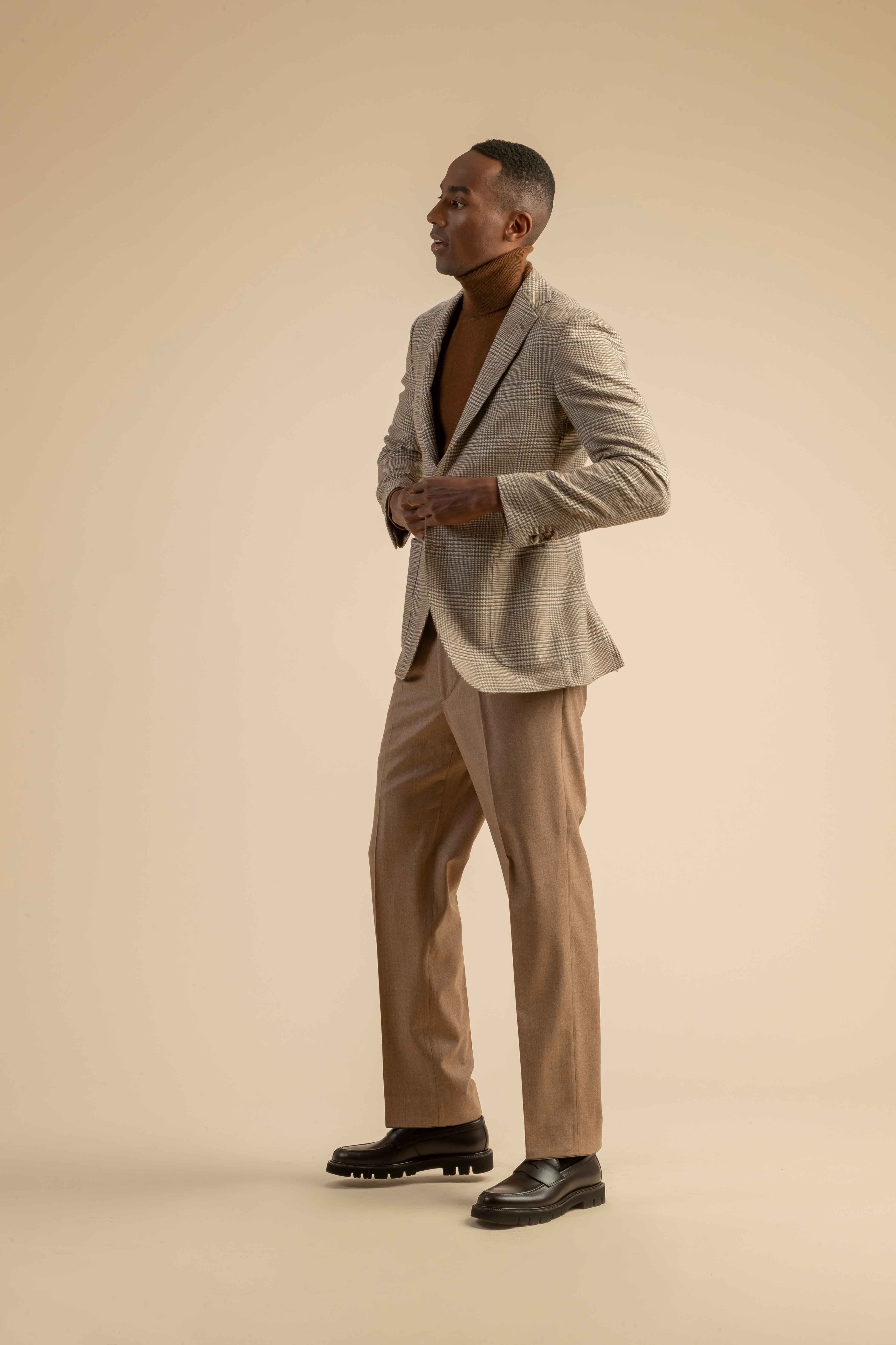 Kariertes Sakko mit Rollkragenpullover und Wollhose aus der Scabal Herbstkollektion Herrenoutfit mit hellbraun kariertem Sakko, braunem Rollkragenpullover und beigefarbener Bundfaltenhose, getragen von einem Model vor beigem Hintergrund.