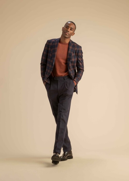 Kariertes Sakko mit rostrotem Pullover aus der Scabal Herbstkollektion Herrenoutfit mit dunkel kariertem Sakko in Blau und Rost, rostrotem Feinstrickpullover und dunkelblauer Stoffhose, getragen von einem Model vor beigem Hintergrund.