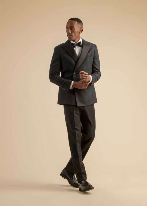 Karo-Smoking mit schwarzem Revers aus der Scabal Herbstkollektion Herren-Smoking mit dunkel kariertem Jackett, schwarzem Revers, weissem Hemd, schwarzer Fliege und klassischer schwarzer Hose, getragen von einem Model vor beigem Hintergrund.