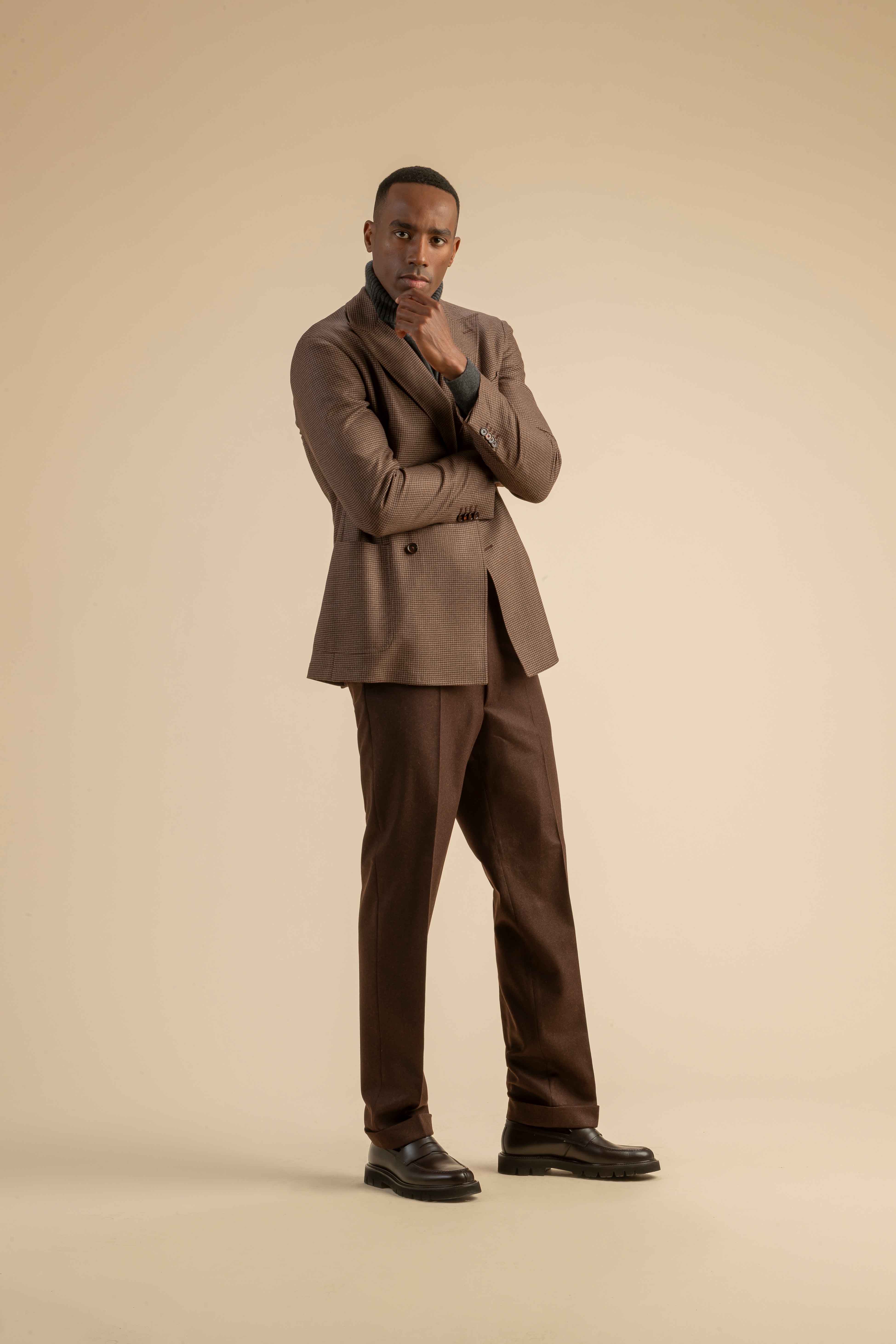 Braunes Karo-Sakko mit Rollkragenpullover aus der Scabal Herbstkollektion Herrenoutfit mit braunem Doppelreiher-Sakko im Mini-Karo, kombiniert mit dunkelgrauem Rollkragenpullover und brauner Bundfaltenhose, getragen von einem Model vor beigem Hintergrund.