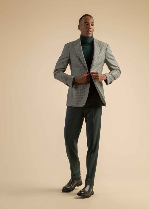 Graues Karo-Sakko mit grünem Rollkragenpullover – eleganter Herbstlook von Scabal Herrenoutfit mit grauem Karo-Sakko, dunkelgrünem Rollkragenpullover und grüner Stoffhose, getragen von einem Model vor beigem Hintergrund.