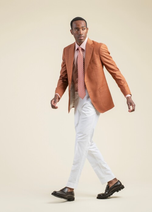 Herrenanzug aus der Scabal Frühling Sommer 2026 Kollektion mit orangefarbenem Sakko aus der Image Linie, kombiniert mit rosa Hemd, gemusterter Krawatte und weisser Hose.